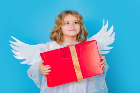 Téléchargez les photos : Kid boy angel with gift box present. Valentine angel. Little cupid child. Kid angel with angels wings, isolated on blue studio background. Valentines day gift card - en image libre de droit