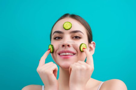 Téléchargez les photos : Woman with facial mask holds slices of cucumber, over blue background. Beautiful young woman with facial mask with cucumber. Skin care and treatment, spa, natural beauty and cosmetology - en image libre de droit