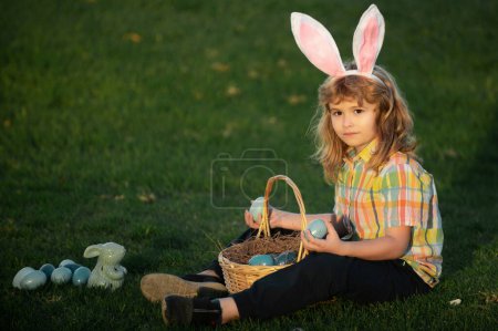 Foto de Conejito con orejas de conejo. Niño niño cazando huevos de Pascua en el césped de primavera que pone en la hierba - Imagen libre de derechos