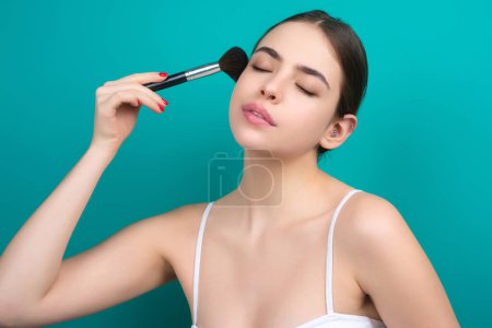 Téléchargez les photos : Young woman applying foundation powder or blush with makeup brush. Facial treatment, perfect skin, natural make up, facial beauty. Isolated on studio background. Applying makeup - en image libre de droit