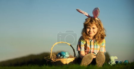 Foto de Wide photo banner for website header design. Cute bunny child boy with rabbit ears. Children hunting easter eggs on sky background with copy space - Imagen libre de derechos
