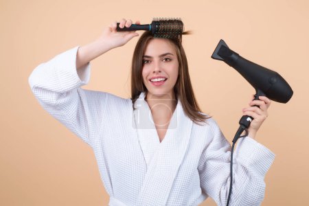 Téléchargez les photos : Woman in bathrobe combing hair, drying hairs with hairdryer. Portrait of female model with a comb brushing hair. Girl with hair brush and blow dryer. Hair care and beauty. Morning routine - en image libre de droit