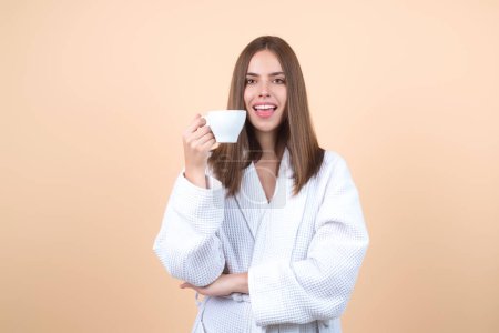 Photo for Morning coffee. Happy young woman with cup of coffee. Smiling girl drinking coffee isolated on beige studio background. Pretty woman in bathrobe with cup of hot coffee - Royalty Free Image