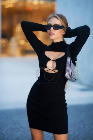 Téléchargez les photos : Modèle de mode en lunettes de soleil de mode, robe sexy noire et sac à main dans la rue. Jeune belle fille sensuelle en vêtements d'été à la mode - en image libre de droit
