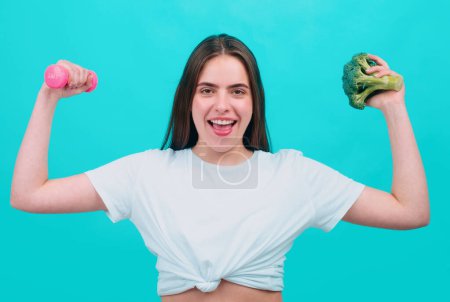 Photo for Portrait of young beautiful woman with broccoli and dumbbell, over blue background. Healthy food, health life - Royalty Free Image