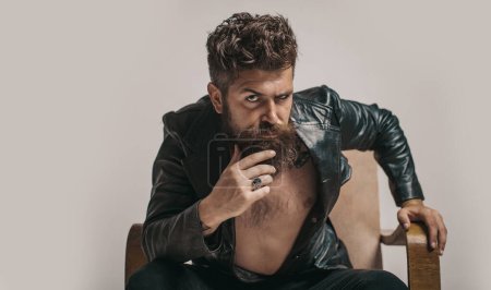 Photo for Brutal bearded fashionable man with serious face sitting in chair and looking at camera. Macho man Casanova attracting women. Sexy rich shirtless millionaire man with leather jacket - Royalty Free Image