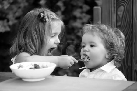 Téléchargez les photos : Soeur nourrissant un petit garçon. La fille nourrit son frère avec une cuillère. Nourriture pour enfants - en image libre de droit