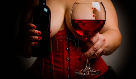 Téléchargez les photos : Femme de taille plus avec une grande belle poitrine tenant un verre avec du vin et une bouteille de vin. Sexy femme chaude avec de gros seins portant corset rouge dans le style victorien. Vin doux et concept femme - en image libre de droit