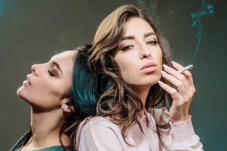 Foto de Dos chicas hermosas jóvenes fumando cigarrillos en un fondo gris. Hábito nocivo y malo de los jóvenes. Las mujeres adicción a la nicotina. No fume, cuide de la salud - Imagen libre de derechos