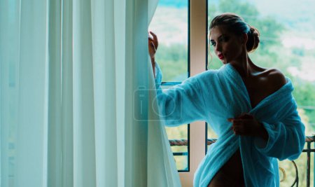 Foto de Hermosa mujer con maquillaje de lujo y peinado en el fondo de la ventana. Sensual mujer joven con albornoz después de tomar una ducha de pie cerca de la ventana y mirando hacia el exterior - Imagen libre de derechos