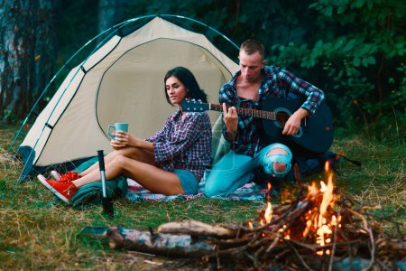 Foto de Jóvenes turistas juntos. Pareja enamorada sentada cerca de carpa turística en el bosque. Pareja enamorada viajando y acampando - Imagen libre de derechos