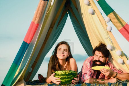 Téléchargez les photos : Couple romantique manger pastèque en tente camping sur fond de nature. Aventure pour les jeunes amis le jour d'été - en image libre de droit