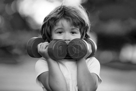 Photo for Cute funny little boy doing exercises with dumbbells in green park. Closeup portrait of sporty child with dumbbells. Happy child boy exercising outdoor. Healthy activities kids lifestyle - Royalty Free Image