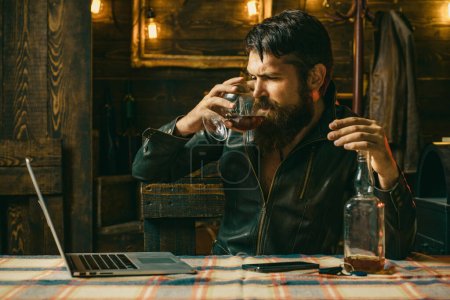 Photo for Drunk bearde sad pensive business man drinking whiskey in front of laptop at loft work place. Man or businessman drinks wiskey at home. Wasted alcoholic businessman drinking whiskey working at laptop - Royalty Free Image