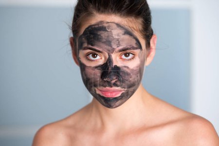 Téléchargez les photos : Traitement du visage. Belle jeune femme avec masque facial en boue de charbon de bois sur le visage. Soin et traitement de la peau. Masque visage en argile noire - en image libre de droit