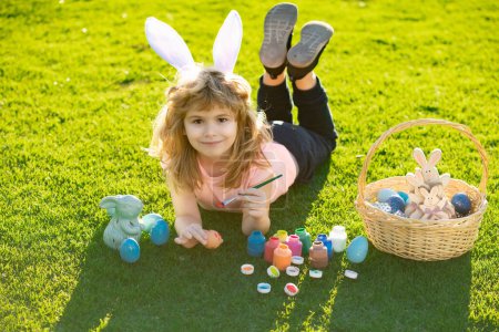 Photo for Child laying on grass in park wit easter eggs. Easter kids boy in bunny ears painting easter eggs outdoor. Cute child in rabbit costume with bunny ears having fun in park - Royalty Free Image