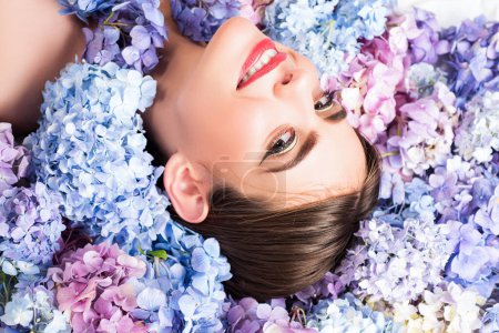 Foto de Young woman face in blooming hydrangea in background. Romantic and love concept. Nature beauty. Woman lying on flowers. Makeup cosmetics and skincare. Girl with hydrangea flowers - Imagen libre de derechos