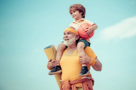 Téléchargez les photos : Grand-père et petit-fils avec ballon de basket et tapis de yoga dans les mains. Homme âgé et mignon petit garçon exerçant sur fond de ciel bleu - isolé - en image libre de droit