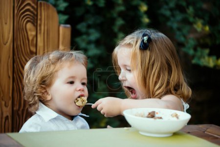 Téléchargez les photos : Soeur nourrissant un petit garçon. La fille nourrit son frère avec une cuillère. Nourriture pour enfants - en image libre de droit