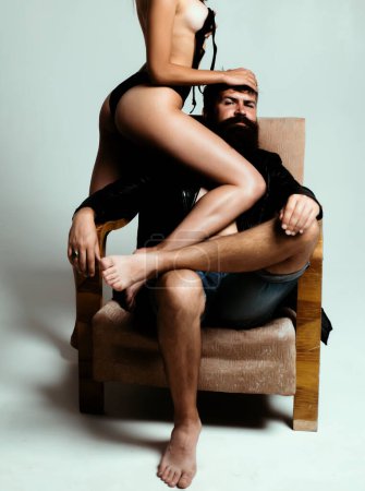 Photo for Brutal bearded man sitting at chair and his sexy naked girl with slim body standing near. Desire and passion of loving couple. Attractive brunette tempting her hipster boyfriend - Royalty Free Image