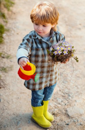 Téléchargez les photos : Heureux petit fermier qui s'amuse sur le champ de printemps. Enfant travaillant dans le jardin près du jardin de fleurs. Portrait de jardinier enfant portant récolté à la ferme - en image libre de droit