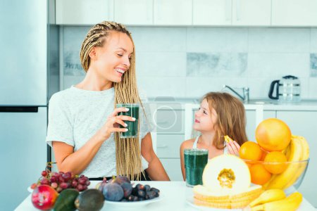 Photo for Mother and daughter drinking green smoothie in a white kitchen. Family mother and child daughter blended green and orange smoothie with healthy fruits ingredients. Detox smoothie and healthy food. - Royalty Free Image