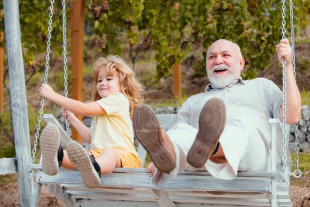 Téléchargez les photos : Excited amazed grandson child and grandfather swinging in summer garden. Granddad and grandchild sitting on swing in park. Weekend with granddad - en image libre de droit