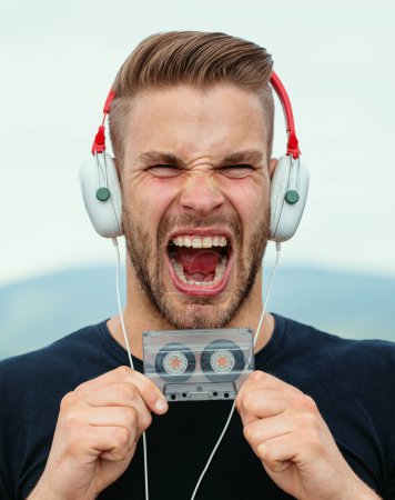 Photo for Excited Man listening music in headphones with cassette. Emotional screaming portrait guy. Young bearded good-looking casual man with and earphones in the style of the 90s, with a retro cassetteplayer - Royalty Free Image