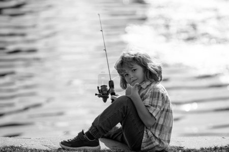 Téléchargez les photos : Pêche des enfants à la rivière. Un jeune pêcheur. Loisirs d'été en plein air. Petit garçon pêchant au bord de la rivière avec canne - en image libre de droit