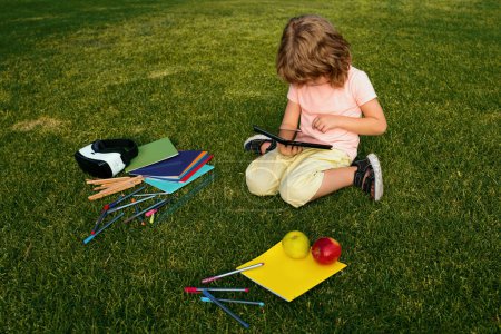 Photo for Kid with tablet sit on grass in park. Kids education, learning and studying. Elementary school students boy doing homework outdoor - Royalty Free Image