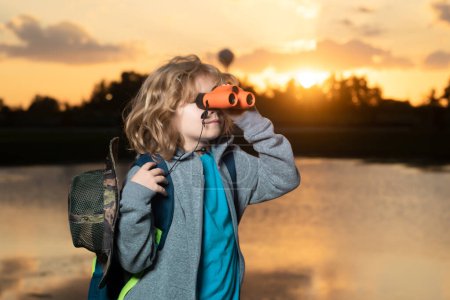 Foto de Young boy looking through binoculars. Cute blond kid with binoculars wearing explorer hat and backpack on nature. Child explorer hiking and adventure - Imagen libre de derechos