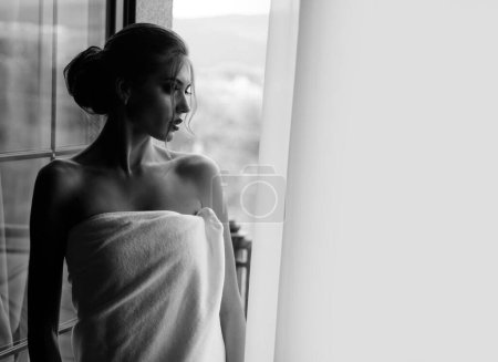 Téléchargez les photos : Sensuelle jeune femme enveloppée dans une serviette après avoir eu une douche debout près de la fenêtre et regardant à l'extérieur. Belle femme avec maquillage de luxe et coiffure à fond de fenêtre - en image libre de droit
