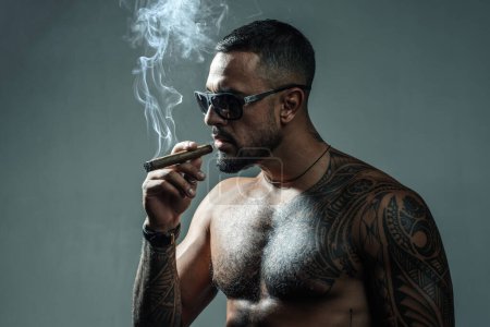 Photo for Strong confident tattooed man in luxury sunglasses smoking an elite tobacco cigar isolated at gray background. Bearded careless man smoke. Attributes of men power, strength and luxury life - Royalty Free Image