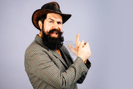 Photo for Young handsome bearded carefree funny man in suit with fingers like guns. Close up photo of fine brunet in hat with imagine gun in hands on isolated background - Royalty Free Image