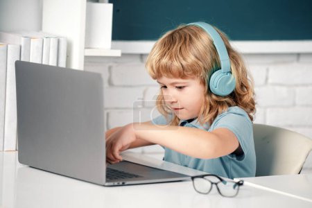 Téléchargez les photos : Petit garçon dans les écouteurs regarder des leçons vidéo sur ordinateur à l'école, enfant dans les écouteurs ont leçon en ligne, en utilisant un ordinateur portable. Programmation géniale pour enfants - en image libre de droit