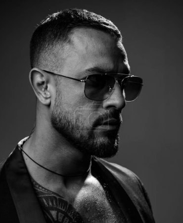 Photo for Sexy bearded tattooed man wearing sunglasses and dramatic face studio photo. Strong muscular man representative of men style and fashion. Luxury lifestyle concept - Royalty Free Image