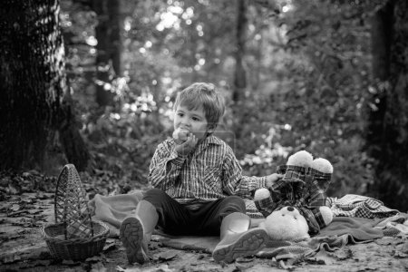 Foto de Divertido caminar en el parque otoñal. Niño sosteniendo un juguete de oso de peluche y comiendo manzana dulce. Concepto de temporada de otoño. Niño bonito relajarse en belleza otoño paisaje fondo - Imagen libre de derechos