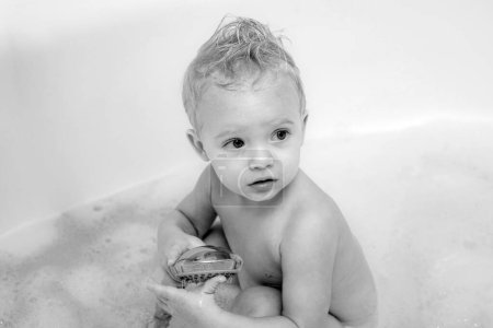 Foto de Ojos azules niño en el cubo de baño mirando a la cámara y sonriendo. Pequeño niño rubio dulce lindo se baña en un baño con espuma y burbujas de jabón - Imagen libre de derechos