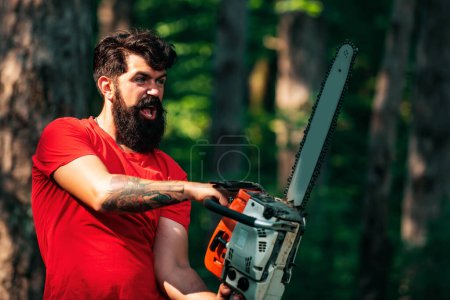 Photo for Lumberjack on serious face carries chainsaw. Woodcutter with chainsaw on sawmill. A handsome young man with a beard carries a tree. Harvest of timber - Royalty Free Image