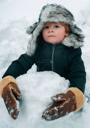 Téléchargez les photos : Enfant mignon portrait d'hiver. Portrait extérieur du petit fils par temps froid et ensoleillé en hiver dans le parc. Rendre le bonhomme de neige et l'hiver amusant pour les enfants. Bonne période hivernale - en image libre de droit