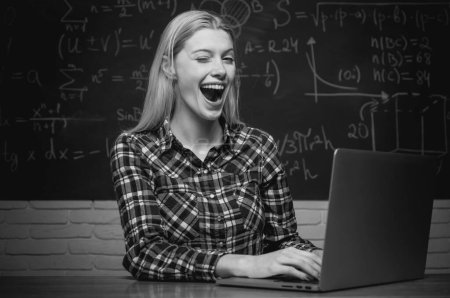 Foto de Female student looking at camera. Cheerful smiling girl student at the blackboard. College tutor. Student sitting at table and writing on notebook. - Imagen libre de derechos