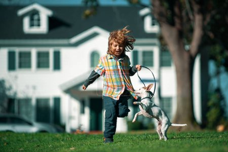 Foto de Perro con correa corriendo con manillar. Niño sobre un fondo de césped verde patio trasero en un día soleado verano. Pequeño cachorro persiguiendo bebé - Imagen libre de derechos