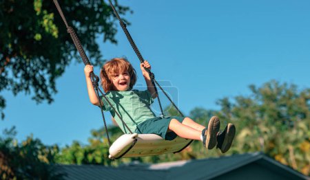 Foto de Columpio infantil al aire libre. Un chico jugando en el patio trasero. Feliz lindo niño balanceo y divertirse saludable actividad de vacaciones de verano - Imagen libre de derechos
