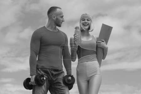 Téléchargez les photos : Jeune couple amoureux d'entraînement en plein air. Entraîneur de sport musculaire avec une jeune femme souriante et joyeuse tenant un tapis de yoga sur fond de ciel nuageux. Couple faisant des activités sportives - en image libre de droit