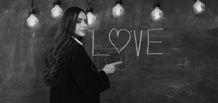 Photo for Students love. Portrait of girl teenager in love. Young cute teenage girl in classroom at blackboard. Young attractive teacher pointing at chalkboard with love text - Royalty Free Image