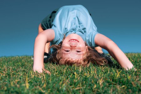 Téléchargez les photos : Portrait d'un enfant garçon souriant heureux jouant à l'envers sur le champ d'herbe en plein air. Un enfant qui rit. Expressions faciales expressives - en image libre de droit