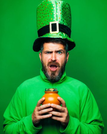 Téléchargez les photos : Homme barbu vêtu d'un costume de lutin isolé sur fond vert. Homme barbu célébrant la Saint Patricks Day. Leprechaun homme en chapeau vert et sweat à capuche tenant pot d'or - en image libre de droit