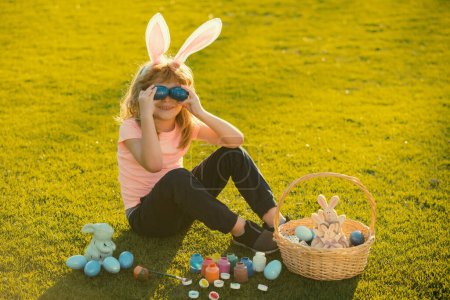 Photo for Child boy with bunny ears cover eyes with eggs on grass background - Royalty Free Image
