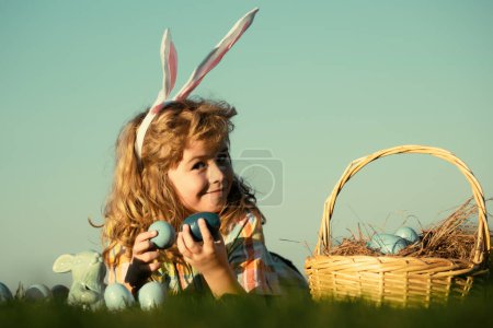 Téléchargez les photos : Des enfants chassant des œufs de Pâques. Petit garçon allongé sur l'herbe et trouvant des œufs de Pâques, sur fond de ciel avec de l'espace de copie. Enfant avec œufs de Pâques et oreilles de lapin, portrait en plein air - en image libre de droit