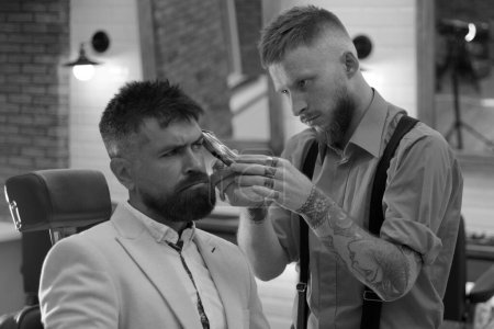 Téléchargez les photos : Coiffeur fait coiffure un homme avec une barbe. Un type brutal dans un salon de coiffure moderne. Lame de rasoir. Homme barbu se faire couper les cheveux par coiffeur et assis dans la chaise au salon de coiffure - en image libre de droit